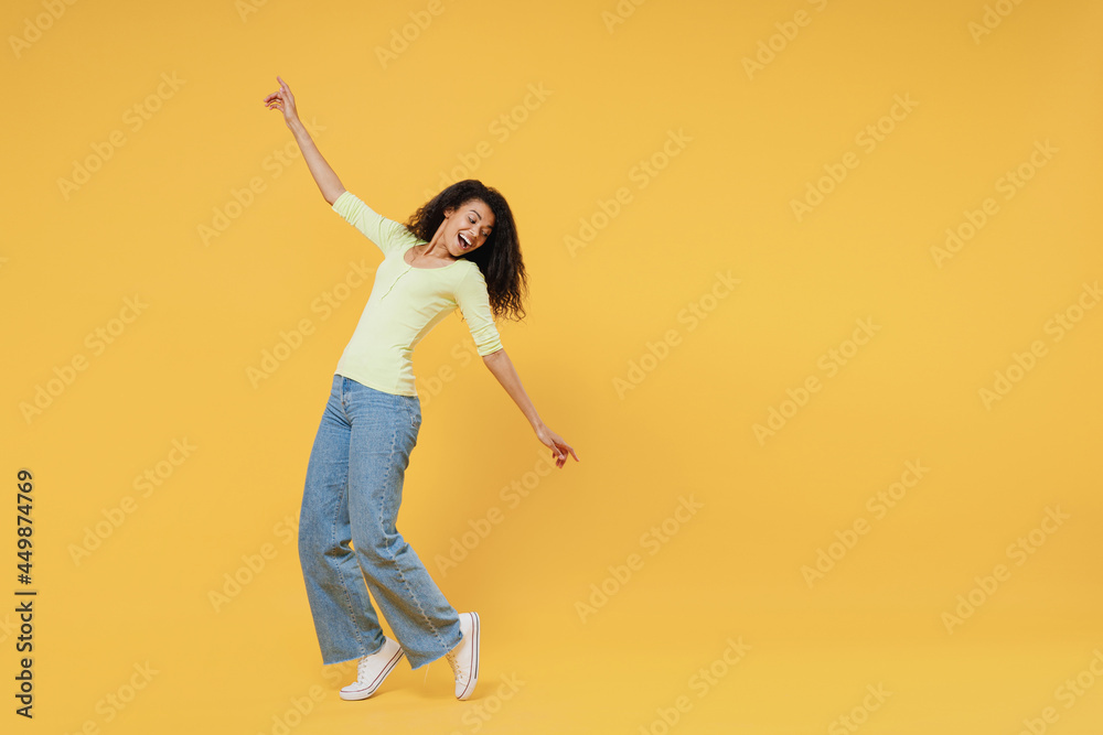 Full size body length smiling overjoyed african american young woman 20s wears green shirt standing on toes dancing lean back have fun spreading hands isolated on yellow background studio portrait