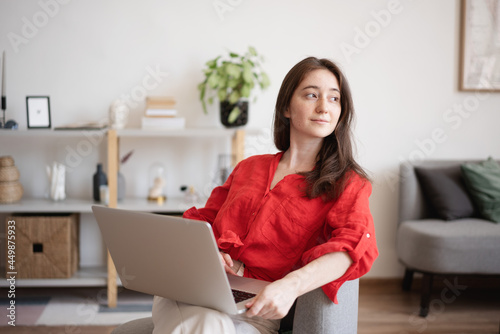 Young happy brunette woman in red blouse working on laptop sitting on chair at home in interior