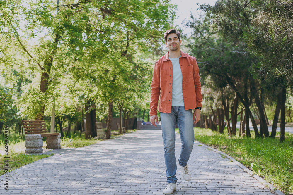 Full length young man 20s wearing orange jacket blue t-shirt walking look camera rest relax in spring green city park go down alley sunshine lawn outdoors on nature. Urban lifestyle leisure concept. Full length young man 20s wearing orange jacket blue t-shirt walking look camera rest relax in spring green city park go down alley sunshine lawn outdoors on nature. Urban lifestyle leisure concept.