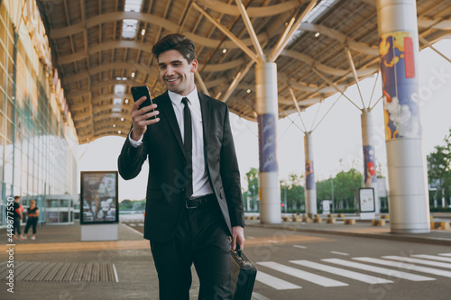 Bottom view fun traveler businessman man in black dinner suit stand outdoorat international airport terminal with attache case hold mobile phone book taxi order hotel Air flight business trip concept