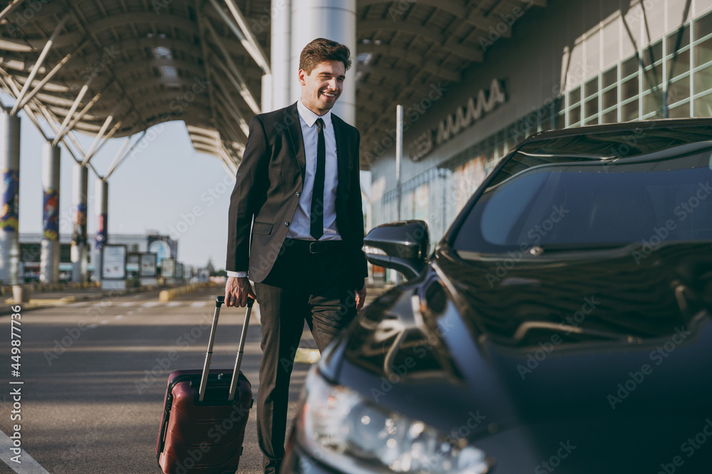 Full body bottom view young traveler businessman young man in black dinner suit going walk outside at international airport terminal with suitcase to car booking taxi Air flight business trip concept Full body bottom view young traveler businessman young man in black dinner suit going walk outside at international airport terminal with suitcase to car booking taxi Air flight business trip concept