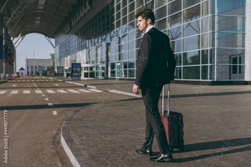 Full length back side view young traveler businessman man in black dinner suit walking going outside at international airport terminal with suitcase valise Air flight business trip lifestyle concept © ViDi Studio