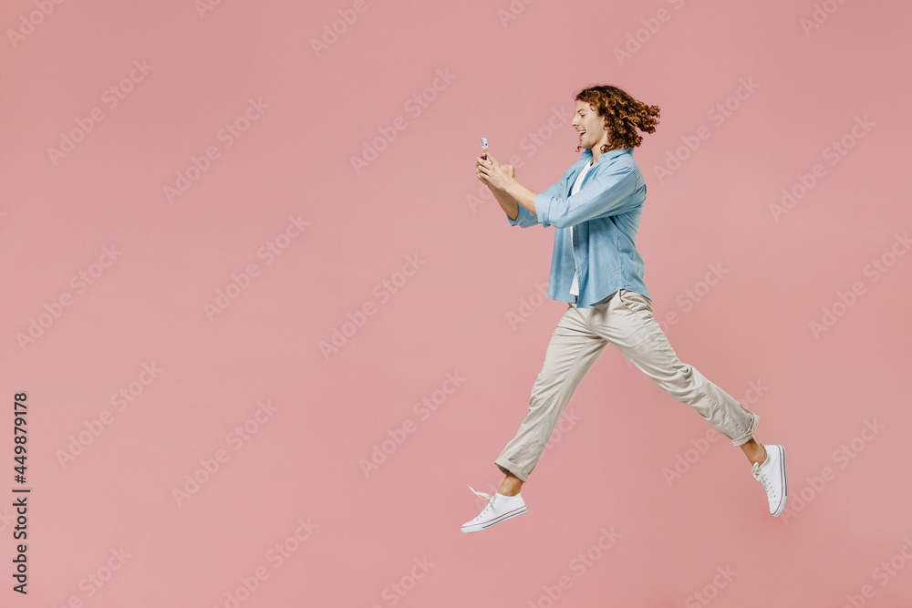 Full size side view young excited happy man 20s with long curly hair wear blue shirt white t-shirt using hold in hand mobile cell phone jump high isolated on pastel plain pink color background studio