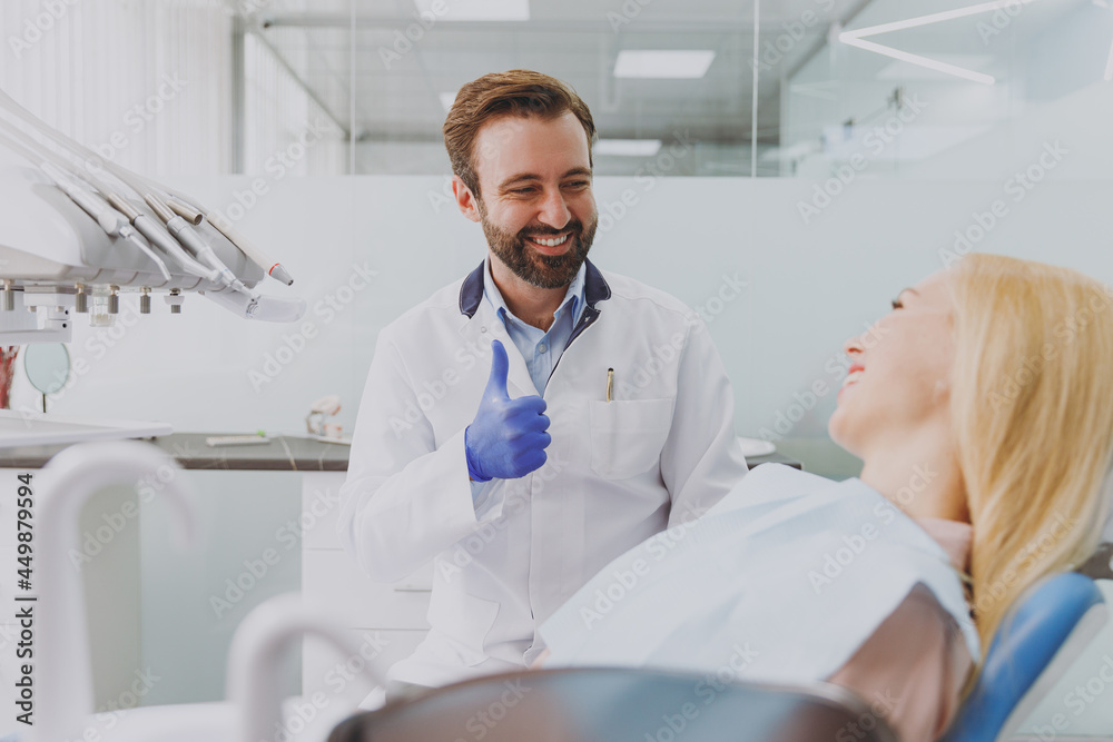 Obraz premium Young smiling fun man dentist doctor in white gown show thumb up gesture consulting patient woman sitting at dentist office chair indoor cabinet near stomatologist Healthcare enamel aesthetreatment