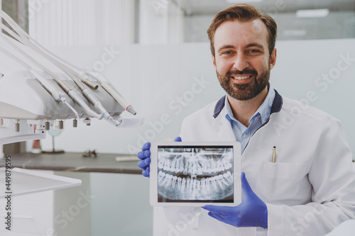 Young fun smiling happy professional orthodontist stomatologist cauasian doctor man 20s wearing white gown show jaw bite teeth x-ray at dentist office indoor cabinet. Healthcare aesthetic treatment photo