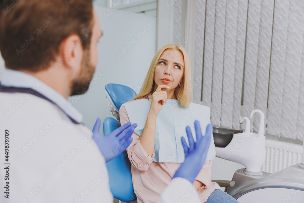 Young man dentist doctor wear white gown talk speak with patient consult minded wistful woman prop up chin sit at dentist office chair indoor cabinet near stomatologist Healthcare enamel treatment