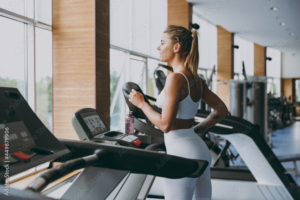 Side profile view young skinny strong sporty athletic sportswoman woman in white sportswear warm up training jogging running on a treadmill in gym indoors. Workout sport motivation lifestyle concept. Side profile view young skinny strong sporty athletic sportswoman woman in white sportswear warm up training jogging running on a treadmill in gym indoors. Workout sport motivation lifestyle concept.