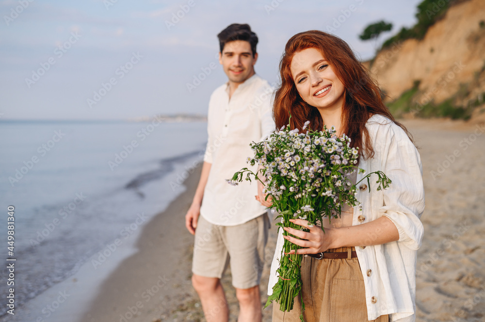 Smiling young couple two family man woman in white clothes hold flowers bouquet hand like follow me walk stroll together at sunrise over sea beach ocean outdoor seaside in summer day sunset evening Smiling young couple two family man woman in white clothes hold flowers bouquet hand like follow me walk stroll together at sunrise over sea beach ocean outdoor seaside in summer day sunset evening