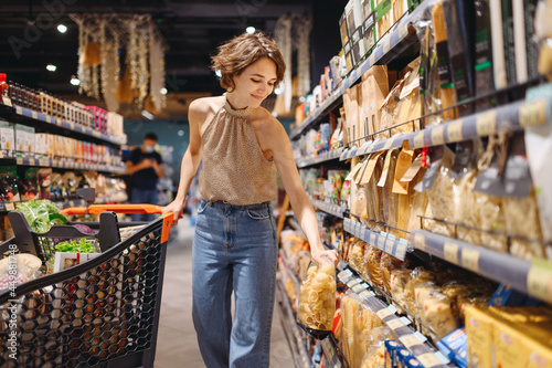 Fototapete Young smiling happy fun caucasian woman 20s wearing casual clothes shopping at supermaket store buy choosing pasta go with grocery cart inside hypermarket
