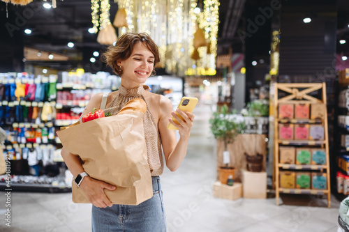 Young woman in casual clothes shopping at supermaket store with craft paper package with groceries using mobile cell phone browsing internet look for recipe inside hypermarket Gastronomy food concept