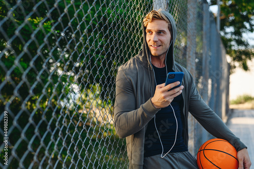 Young fun smiling serious minded sportsman man 20 in grey sportswear hood headphones listen to music use mobile cell phone hold ball at basketball playground court Outdoor courtyard sports concept
