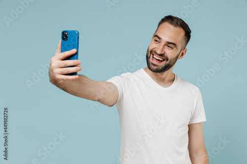 Young smiling fun happy man in white t-shirt doing selfie shot on mobile cell phone post photo on social network isolated on plain pastel light blue color background studio. People lifestyle concept.
