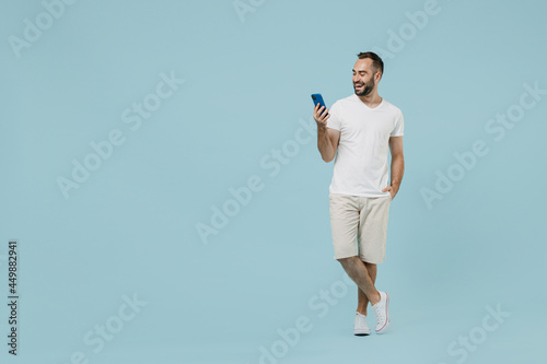 Full length young smiling friendly happy man 20s wearing casual white t-shirt looking camera using mobile cell phone chat onine isolated on plain pastel light blue color background studio portrait