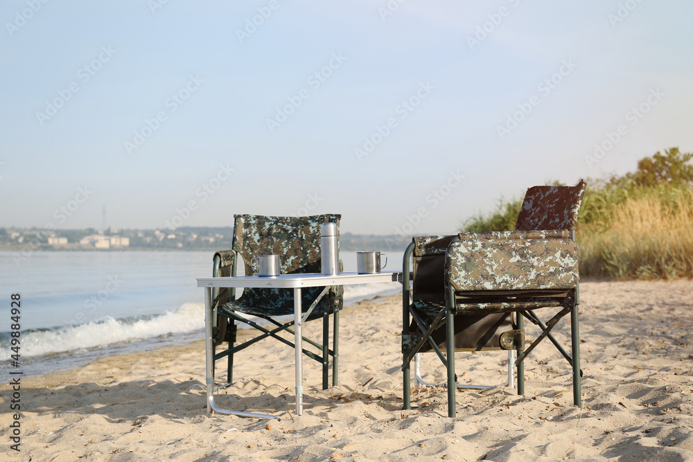 Camouflage fishing chairs and table with metal cups on sandy beach near river Camouflage fishing chairs and table with metal cups on sandy beach near river