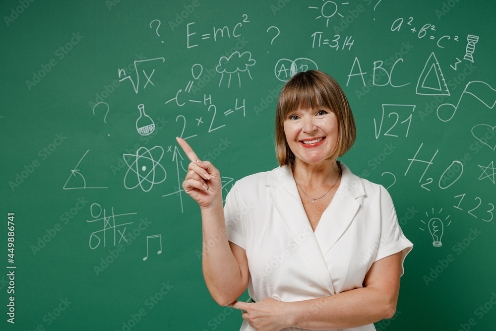 Smiling smart teacher mature elderly senior elegant lady woman 55 wears white shirt point finger above on workspace area copy space mock up isolated on green wall chalk blackboard background studio.