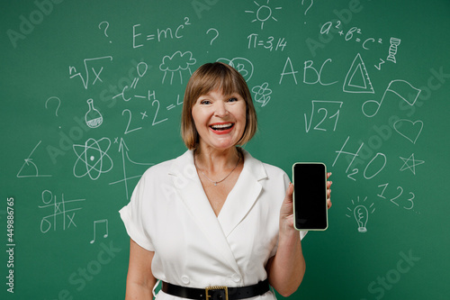 Smiling fun teacher mature elderly senior lady woman 55 wear white shirt hold in hand use mobile cell phone with blank screen workspace area isolated green wall chalk blackboard background studio © ViDi Studio