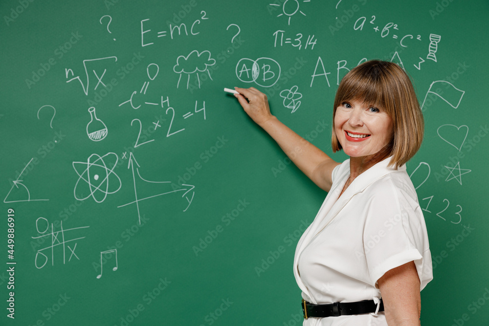 Happy fun teacher mature elderly lady woman 55 wear white shirt hold in hand write point with chalk isolated on green wall blackboard background studio. Education in high school September 1 concept. Happy fun teacher mature elderly lady woman 55 wear white shirt hold in hand write point with chalk isolated on green wall blackboard background studio. Education in high school September 1 concept.