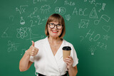 Teacher mature elderly lady woman 55 wear shirt glasses hold takeaway delivery craft paper brown cup coffee to go show thumb up like gesture isolated on green wall chalk blackboard background studio.