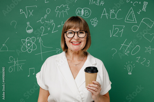 Smiling cheerful clever fun teacher mature elderly lady woman 55 wear shirt glasses hold takeaway delivery craft paper brown cup coffee to go isolated on green wall chalk blackboard background studio. © ViDi Studio