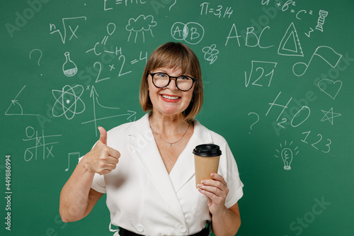 Teacher mature elderly lady woman 55 wear shirt glasses hold takeaway delivery craft paper brown cup coffee to go show thumb up like gesture isolated on green wall chalk blackboard background studio. © ViDi Studio