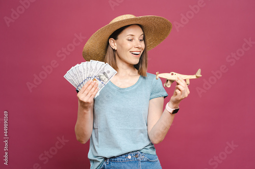 Portrait of a young woman in sunglasses, hat with money and wooden airplane on pink background