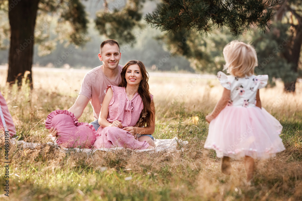 Naklejka premium Mom, dad and little daughter are sitting next to wigwam decoration in the park. Family spending time outdoor in summer, having fun together. Girl are dressed in pink dress
