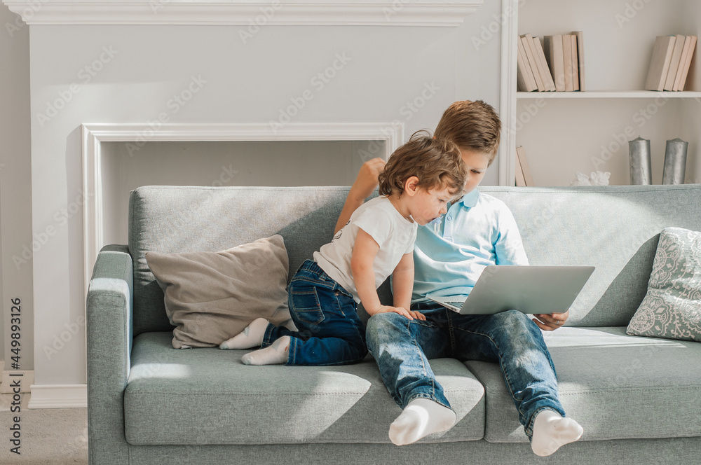 Two little kids siblings seated on sofa in living room holding on lap laptop play online game, watch cartoons spend weekend using electronic device, bad habit, generation addicted with gadgets concept Two little kids siblings seated on sofa in living room holding on lap laptop play online game, watch cartoons spend weekend using electronic device, bad habit, generation addicted with gadgets concept