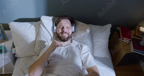 Dreamy attractive young man lying in bed, smiling and listening to music in headphones. Feeling joyful, moving in rhythm, singing. photo