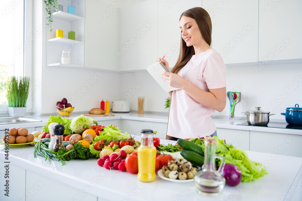 Profile side view portrait of attractive cheery long-haired girl cooking fresh yummy homemade salad writing in light white home kitchen indoors