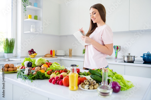 Profile side view portrait of attractive cheery long-haired girl cooking fresh yummy homemade salad writing in light white home kitchen indoors