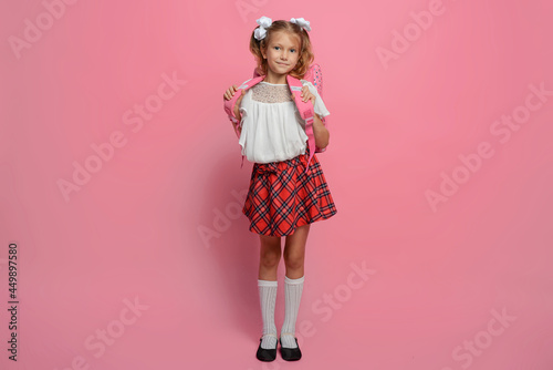 Elementary school girl carries a backpack. Isolated on pink background
