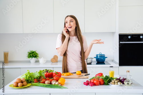 Portrait of attractive cheery dreamy girl cooking tasty fresh dish salad listening talking in light white home kitchen indoors