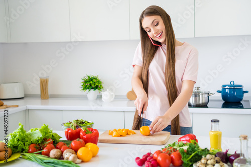 Portrait of attractive cheerful long-haired girl cutting tomato talking on phone at home light white kitchen indoors