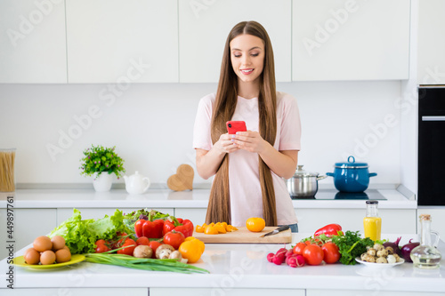 Portrait of attractive cheerful focused girl cooking tasty yummy salad using device chopping tomato in light white home kitchen indoors