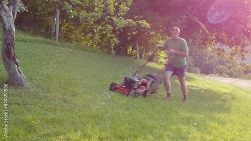Man mowing lawn between fruit trees with red petrol rotary lawnmower machine. Aerial 4k view. photo
