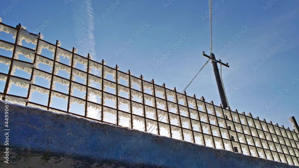 Fototapeta premium Fluffy frost on a metal wire fence against the background of an electric pole and blue sky