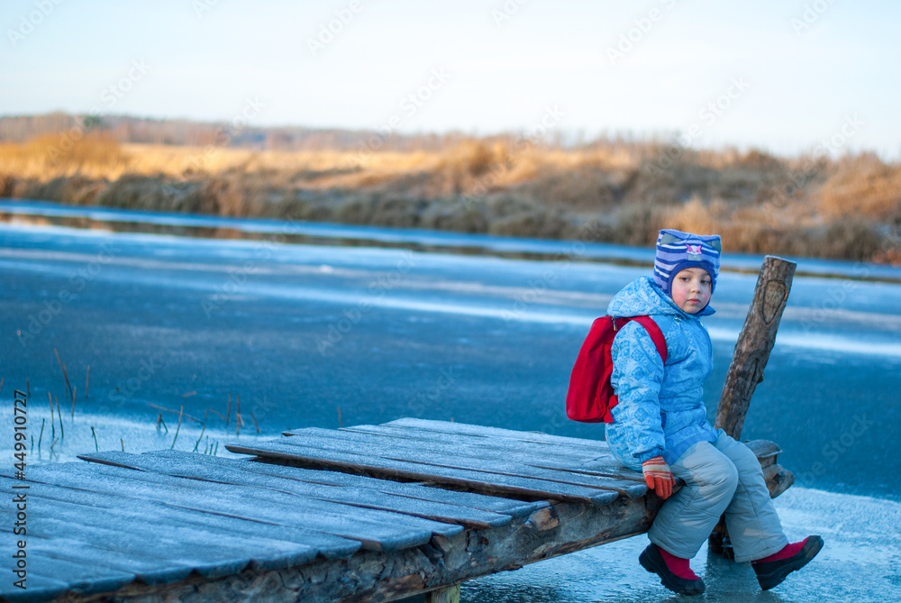 A little girl in a winter blue jacket and with a red backpack sits on a pier above a frozen river and holds her feet above the ice