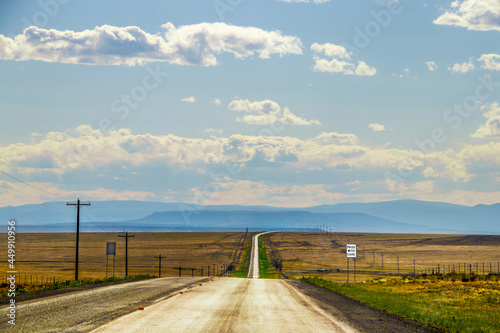 Silver line of two lane highway stretches to horizon with blue layered mountains in the distance and dry fields  and electric lines on each side near dusk with Pass With Care sign.