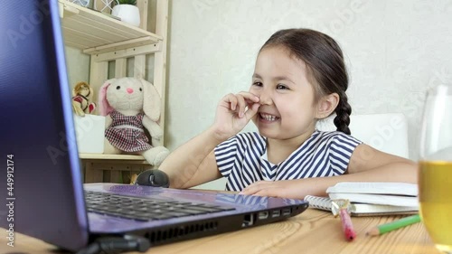 Brown-haired small girl studying at home doing school homework with laptop and picking one's nose. Facial expressions and emotions.