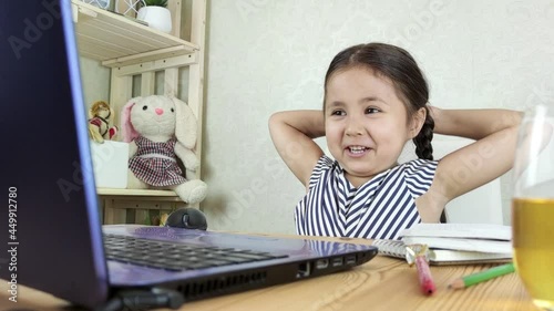 Brown-haired small girl is surfing the Internet sitting at the computer at home. Little 4-year-old girl laughs looking into a laptop. Distance learning online education.