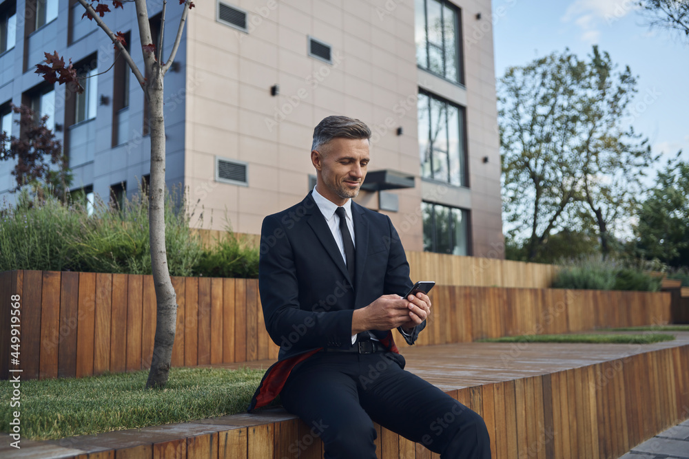 Confident mature businessman using smart phone while sitting outdoors near office building Confident mature businessman using smart phone while sitting outdoors near office building