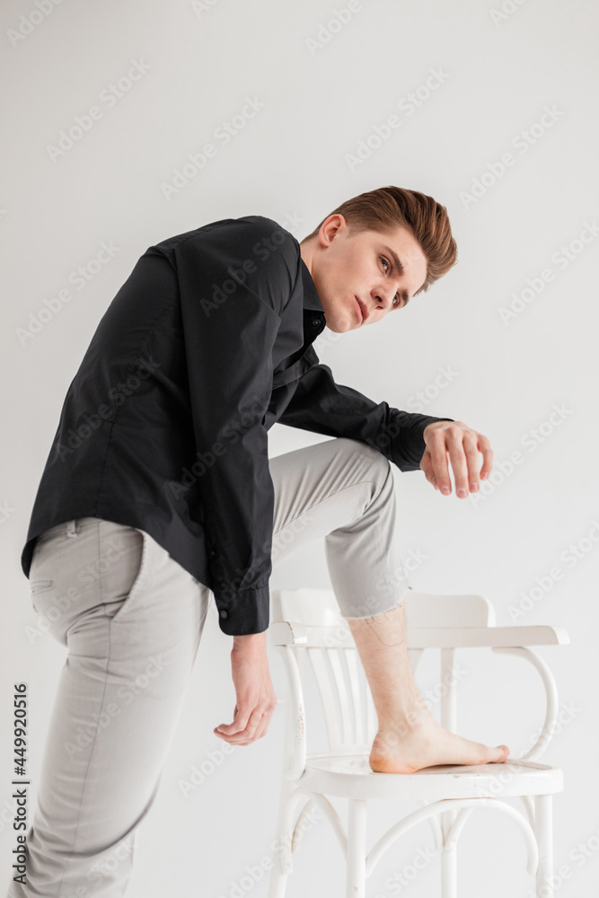 Nice good-looking young man with trendy hairstyle in fashionable black shirt in stylish pants stands near wooden chair on white background indoors. Pretty elegant guy model posing near wall in studio. Nice good-looking young man with trendy hairstyle in fashionable black shirt in stylish pants stands near wooden chair on white background indoors. Pretty elegant guy model posing near wall in studio.
