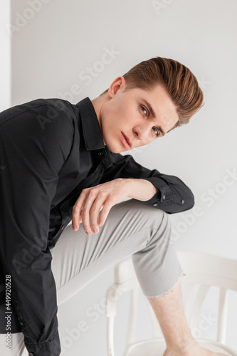 Handsome young man in elegant black shirt in in vintage fashionable pants posing near wooden chair in studio. Attractive guy fashion model in classic stylish clothes near white wall in room.