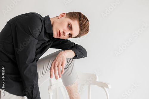 Fashion model good-looking young man with trendy hairstyle in fashionable black shirt in stylish pants stands near chair on white background in studio. Handsome elegant guy posing near wall indoors.
