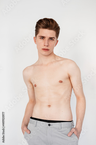 Handsome young man with fashionable hairstyle in gray vintage pants with naked torso stands on white background indoors. Sexy guy fashion model with healthy body posing near white wall in studio.