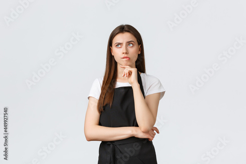 Small business, employees and coffee shop concept. Complicated thougtful saleswoman in black apron thinking. Serious barista making plan, looking up focused while pondering decision photo