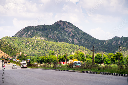 Wide shot of indian highway with trucks, cars, bikes, motorcycle and more on a wide asphalt road with green plants covering hills with clouds of monsoon rains casting shadows showing the beautiful roa