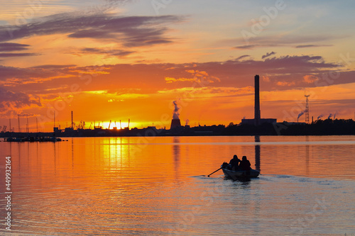 A beautiful sunset on the river, a boat with oars with a soft effect against the background of blast furnace factory pipes and port cranes. Landscape background. The concept of a boat trip.
