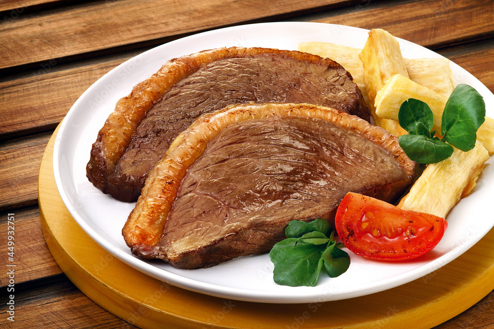 prato com picanha fatiada acompanhada de mandioca frita, tomate e folhas verde sobre fundo de madeira prato com picanha fatiada acompanhada de mandioca frita, tomate e folhas verde sobre fundo de madeira