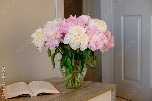 Close up shot of the dresser top with a bouquet of peonies in a glass vase and an open book. Good morning concept. Bedroom with an open door. Copy space for text, background, top view, close up.